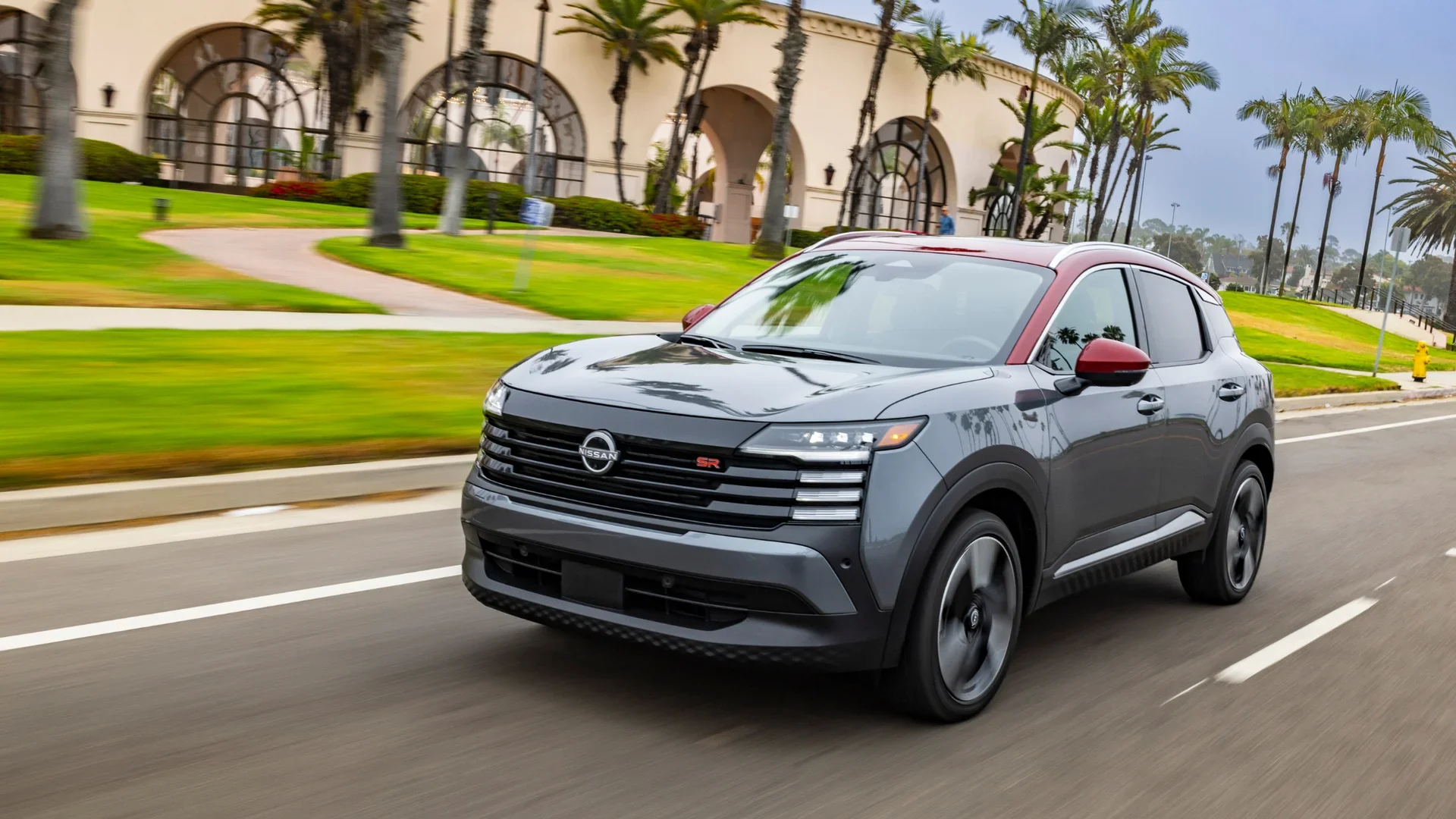 2026 Nissan Kicks® SR in gray with red roof, driving past palm trees and a landscaped urban setting.