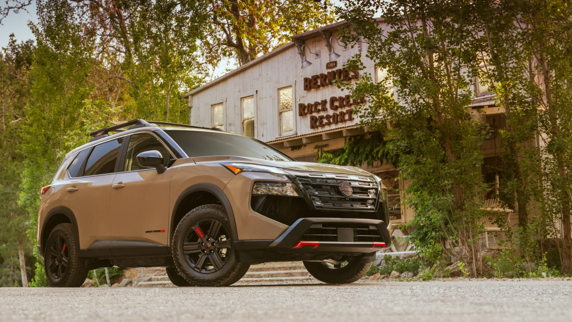 2026 Nissan Rogue® Rock Creek in beige parked outdoors near a rustic mountain resort building scene.
